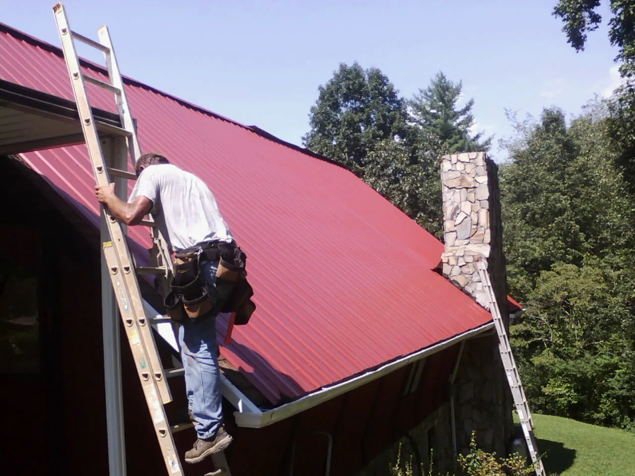 Roofer on a ladder beside a red metal roof and stone chimney — Soffit & Fascia Repair by Roofing Brothers in Buxton
